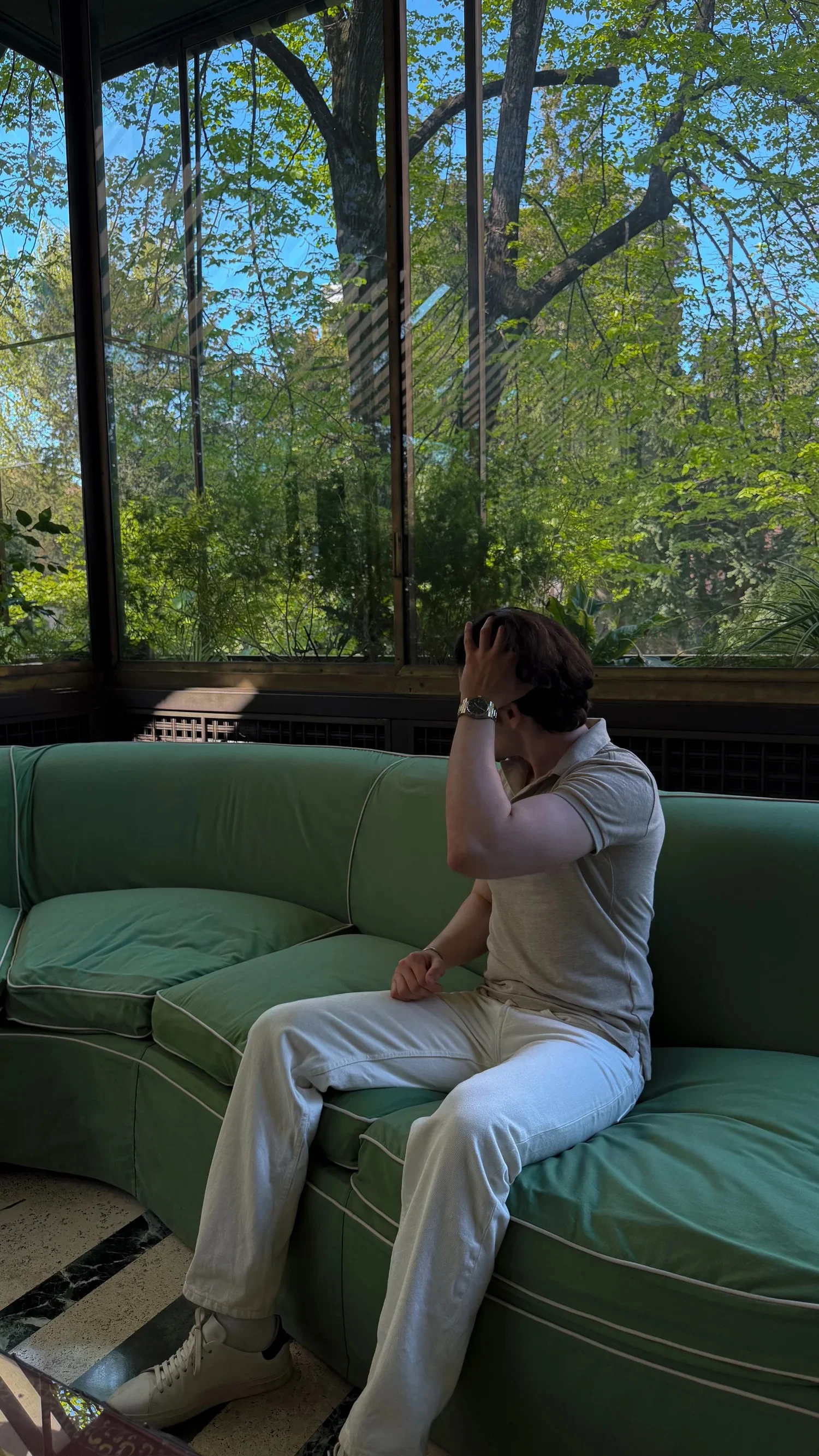 A person seated on a green sofa in a glass-walled garden room.