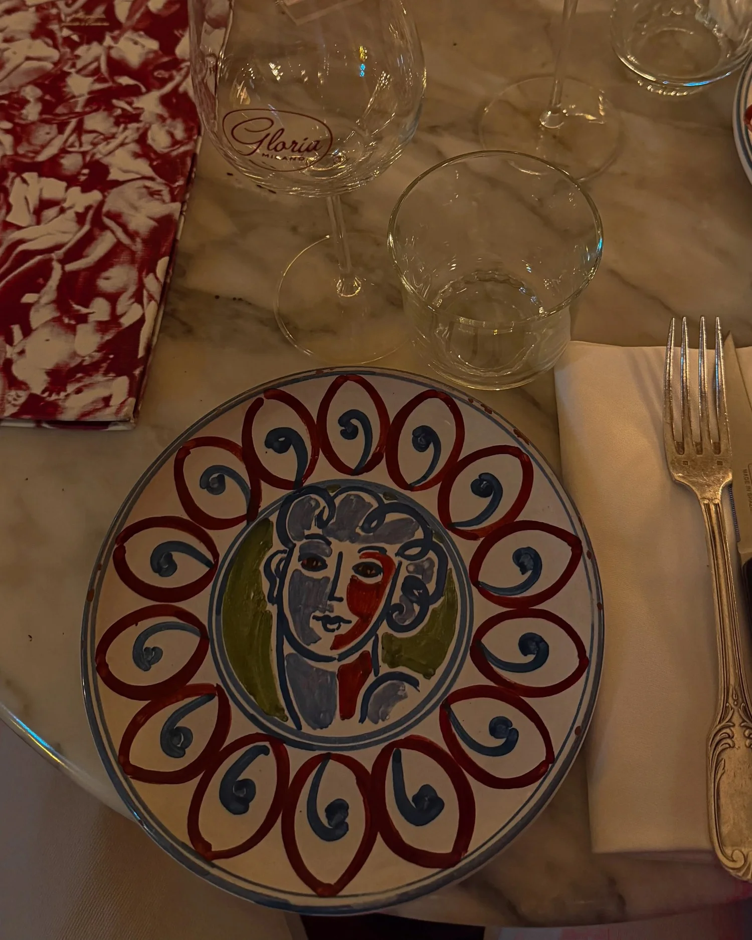 Table setting at Gloria Osteria in Milan with decorative plate, branded wine glass, marble tabletop, and red patterned napkin.