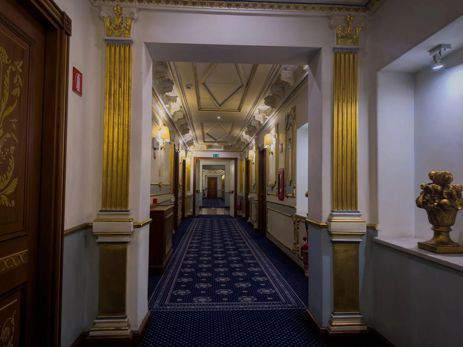 Hotel corridor with patterned blue carpet, gilded trim and decorative columns.