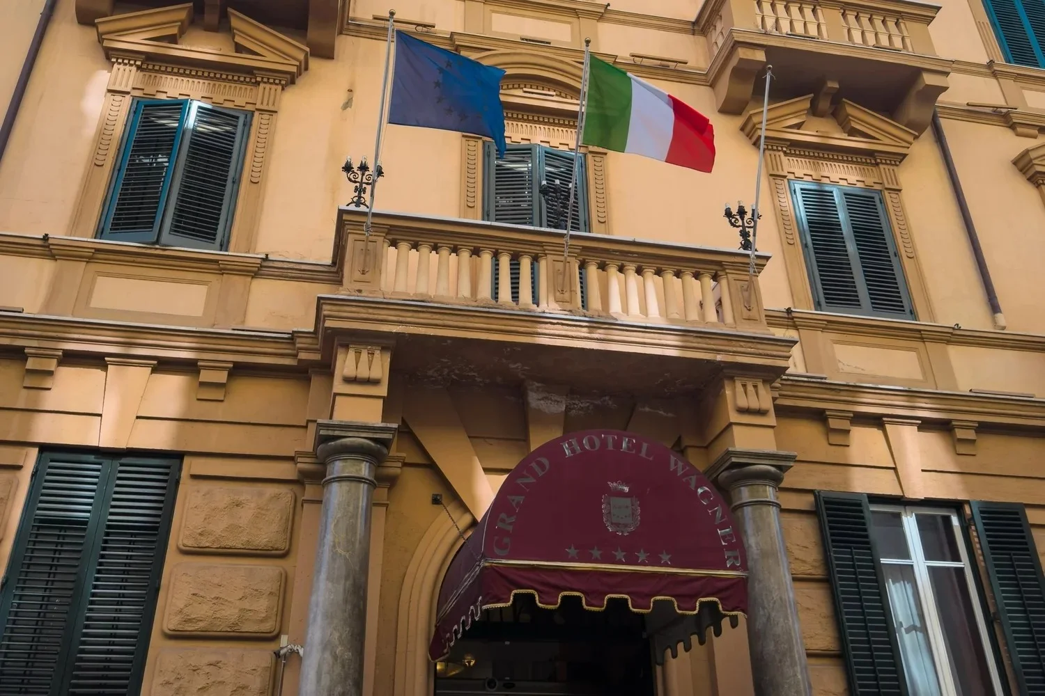 Grand Hotel Wagner entrance with flags, blue shutters and a burgundy canopy.