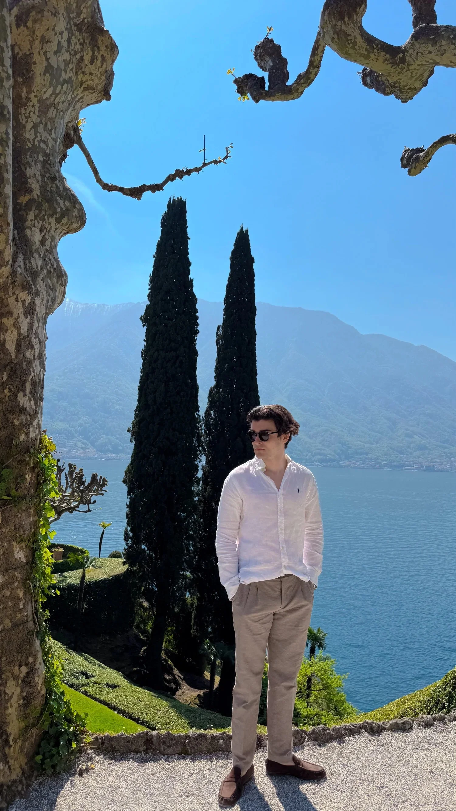 A person standing between trees on a lakeside garden terrace.