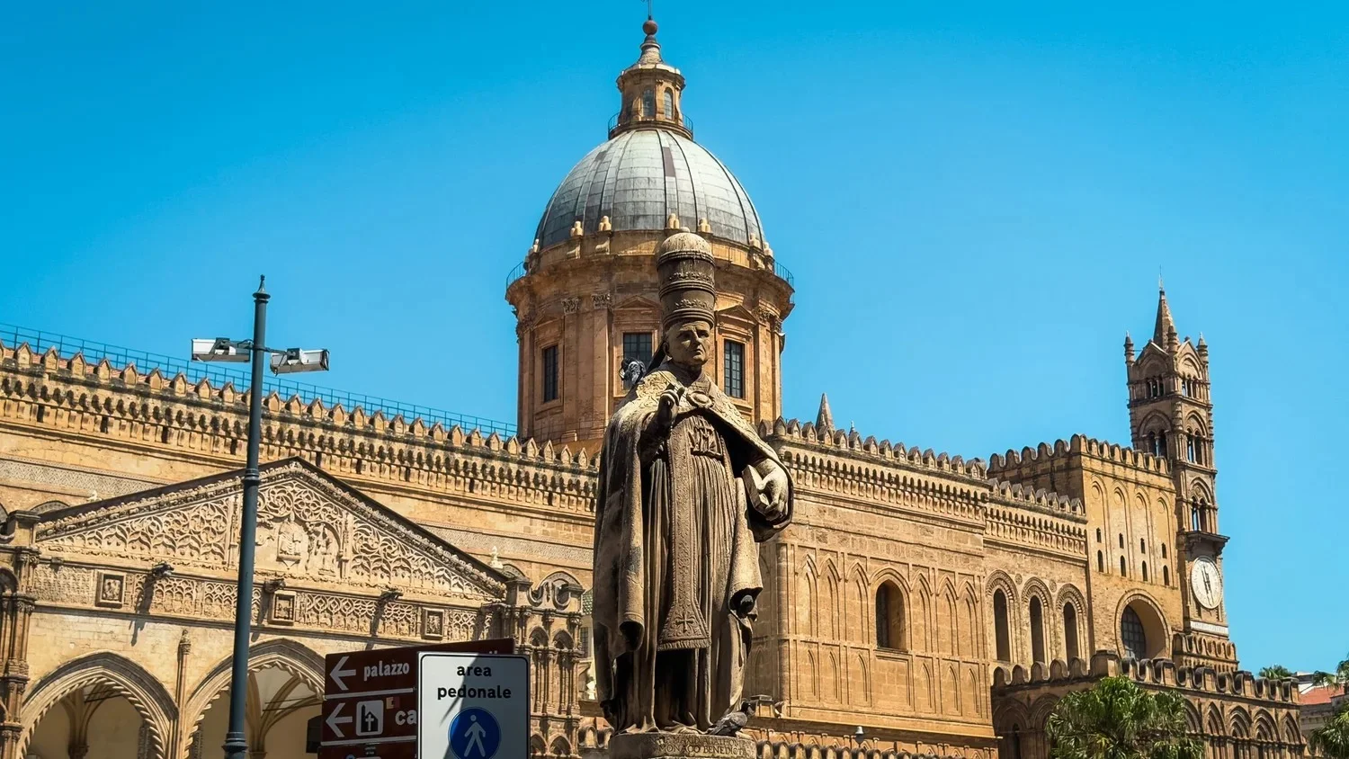 Palermo Cathedral with its dome, crenellated walls and a stone statue in the foreground under a blue sky.