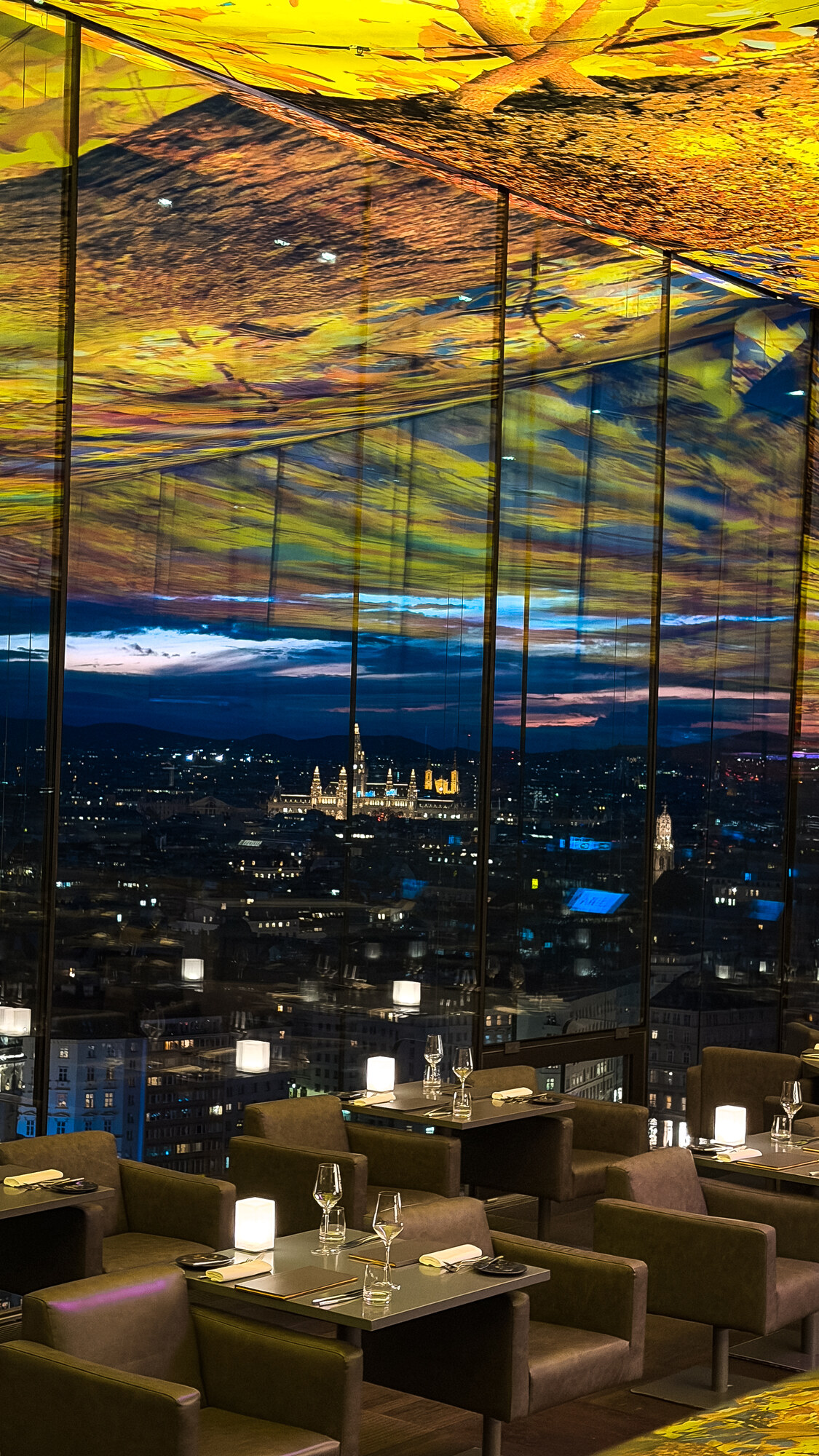 DAS LOFT’s illuminated yellow ceiling reflected in tall windows above lamp-lit tables and Vienna’s evening skyline.