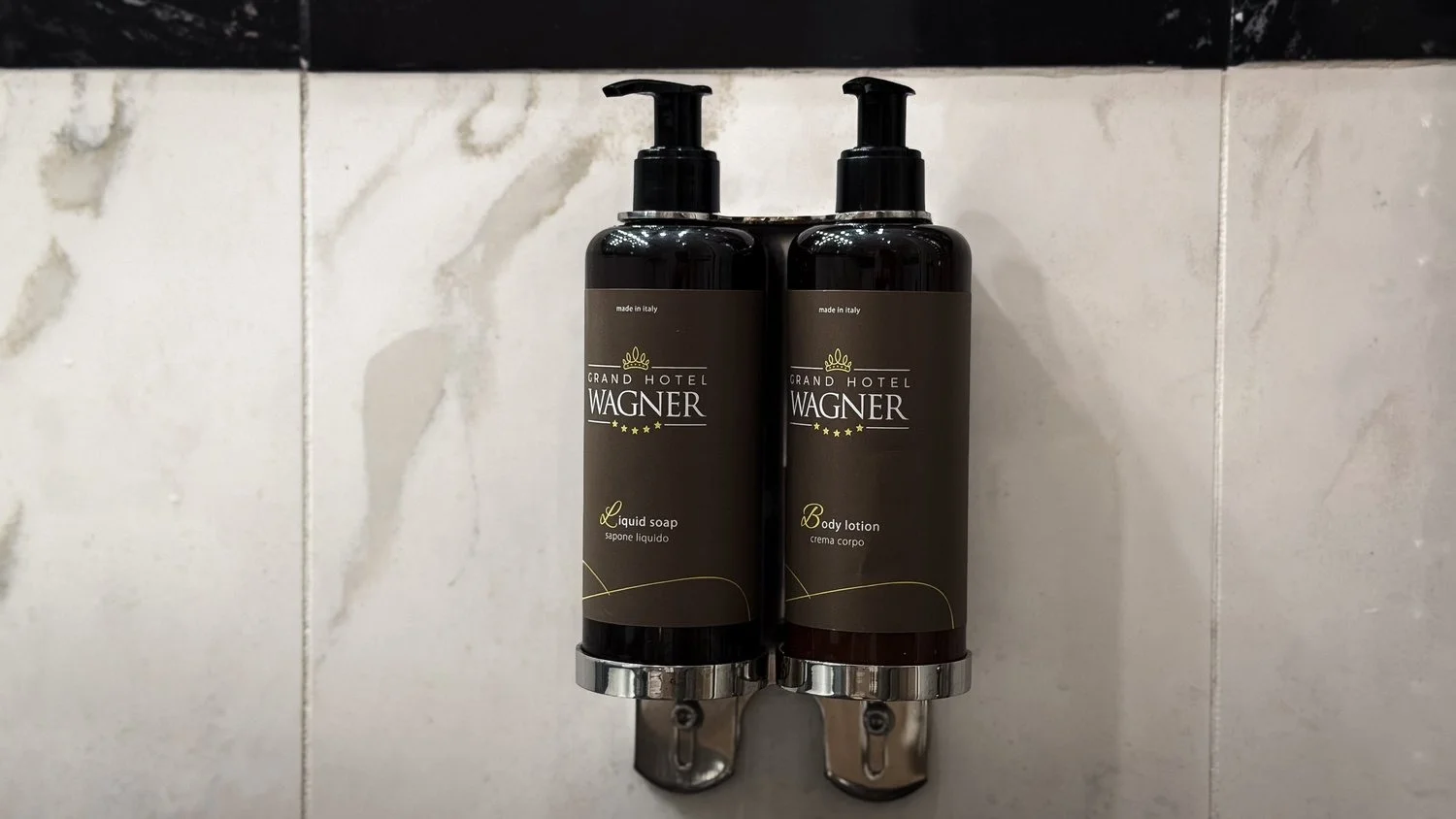 Grand Hotel Wagner toiletries mounted against a marble shower wall.