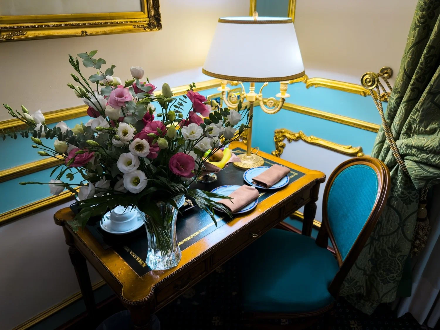 Flowers and a welcome tray on an ornate desk beside a lamp.