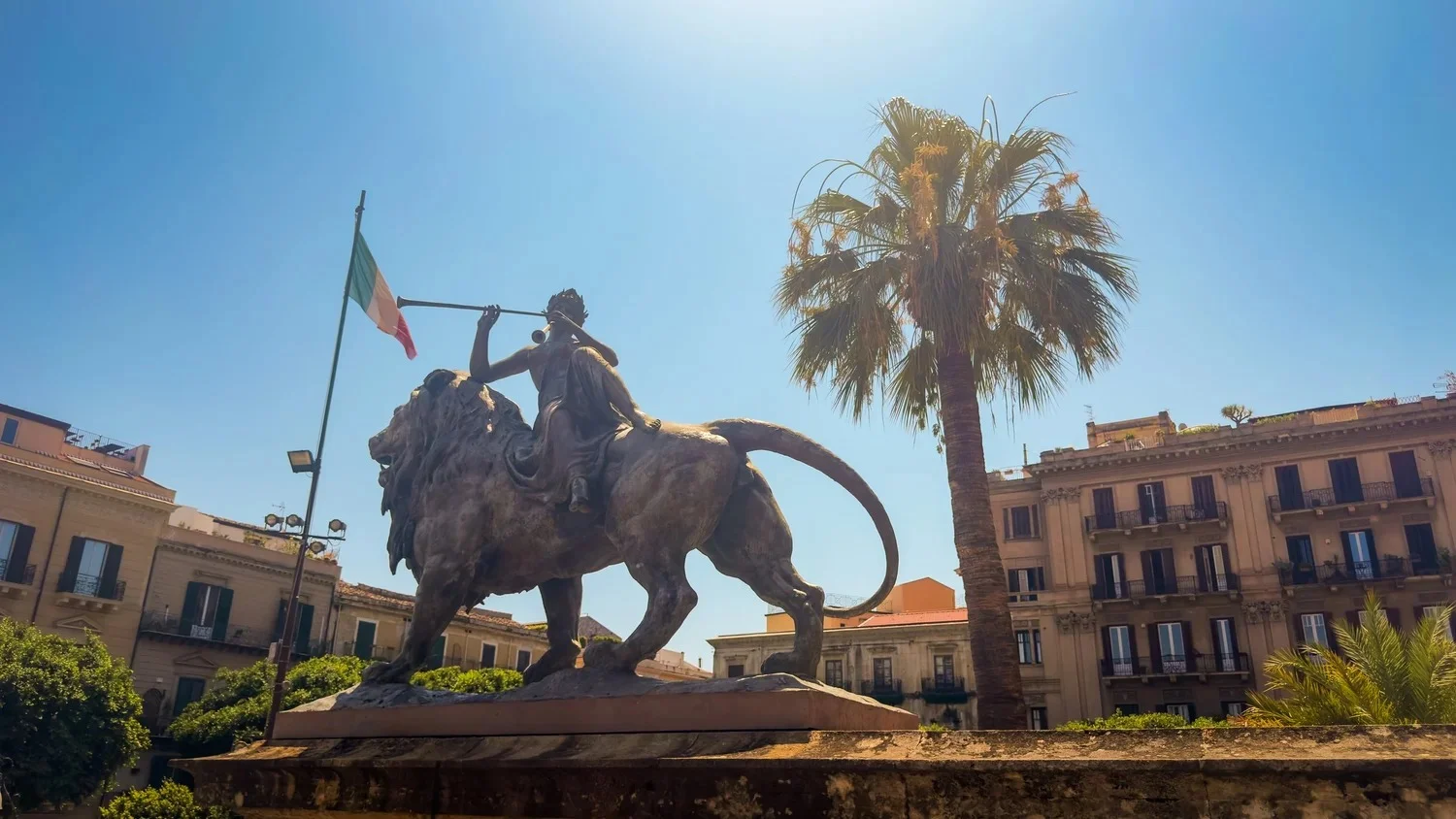 Bronze lion topped by the allegorical figure of La Lirica outside Teatro Massimo in Palermo, with a palm tree and Italian flag behind.
